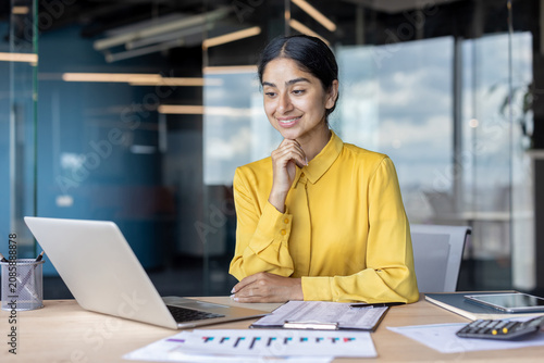 A young Indian woman sits at an office desk and looks at her laptop screen with a smile