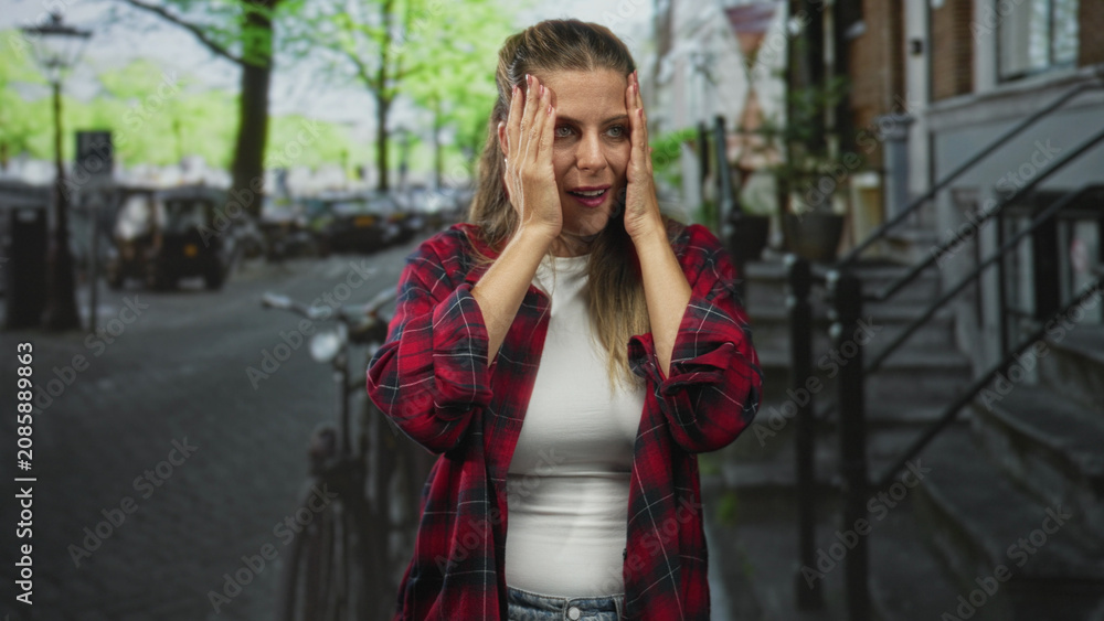 Fototapeta premium Young woman in red plaid shirt with hands raised and wide eyes, standing by a bicycle on a tree lined street; surprise explanation.