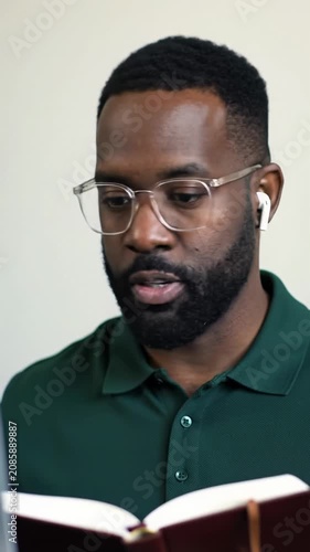 Young adult african-american man, mid-thirties, in dark green polo & glasses, jotting notes during virtual meeting in minimalist home office, soft natural light, dolly in, calm productivity concept