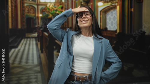 Valokuva Woman wearing glasses flips hair with right hand, left hand in pocket, white crop top and belt visible while standing by pews and tiled aisle in ornate building; confidence