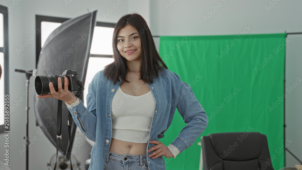 Fototapeta premium Young woman holding camera with hand on hip in a studio with green screen and lighting; confidence creativity.
