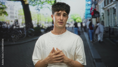 Fotografie Man in white shirt standing on a city street with hands clasped near chest and hesitant speaking gesture, worried expression; concern