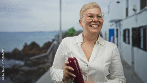 Bild auf Leinwand Woman holding a juice bottle by a seaside building, smiling and clutching the bottle with both hands; health vitality joy