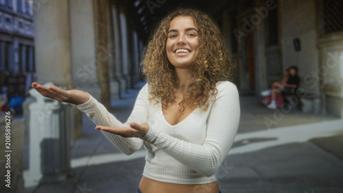 Foto Young woman with curly hair presenting palms up, smiling, showing navel piercing and white cropped sweater amid street colonnade and stone arches; warm friendly joy