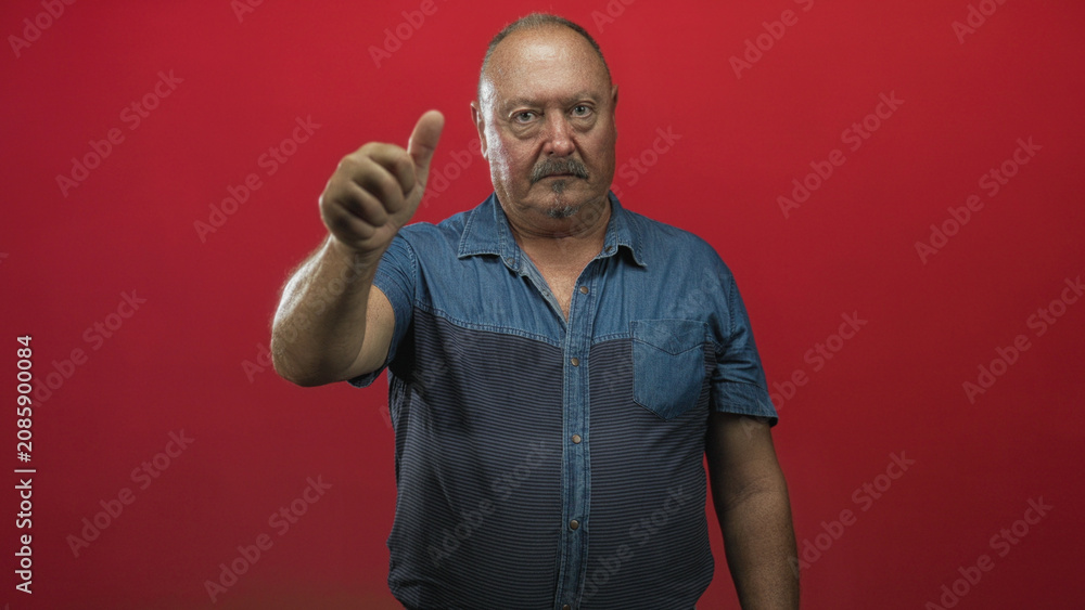 Fototapeta premium Man showing thumbs up with right hand and visible thumb, wearing denim shirt in studio red backdrop; confidence support encouragement.