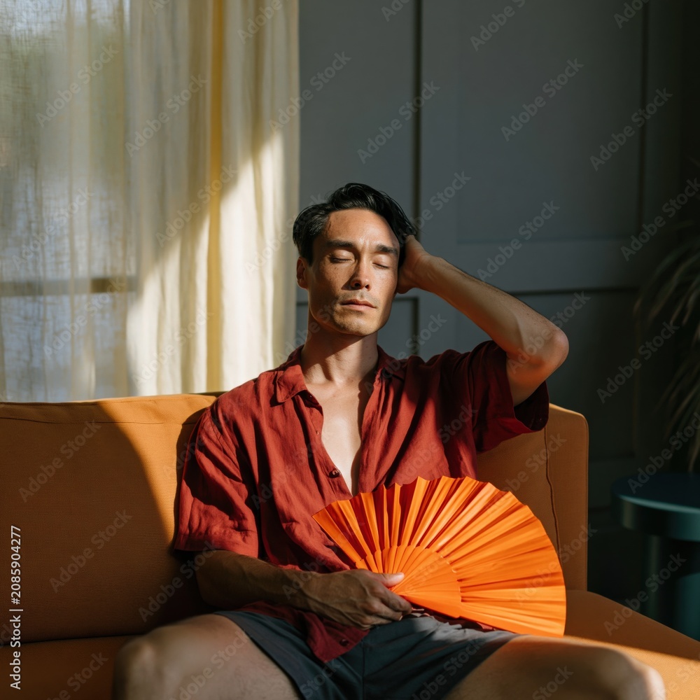 Obraz premium Man sitting on sofa fanning himself with orange hand fan in sunlight wearing red shirt for summer lifestyle and editorial background