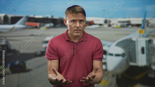 Konstfotografi Man with open palms and pleading gesture at an airport gate, standing before jetbridge and airliner while wearing a red polo shirt; travel delay frustration