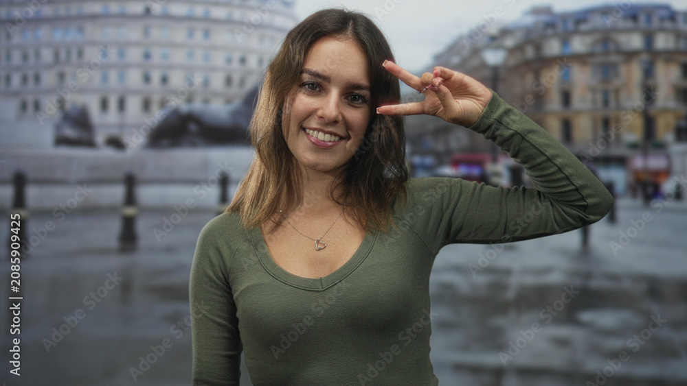 Fototapeta premium Young hispanic woman smiling and making peace sign near face on wet city street, wearing green shirt and heart necklace; youthful joy.