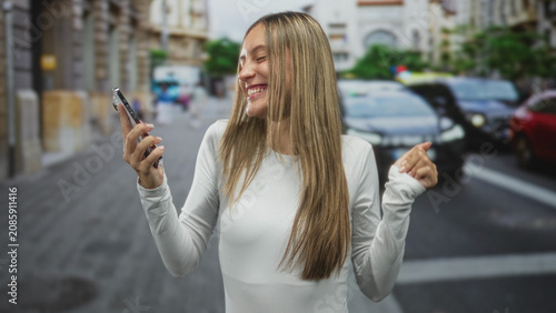 Foto Young woman holds smartphone and pumps fist on city street while laughing and dancing; joy youthful energy