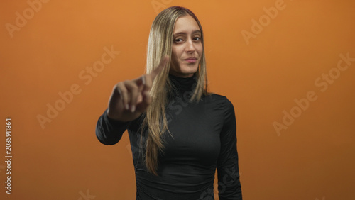 Fotografía Young woman pointing index finger at camera in orange studio backdrop wearing black turtleneck; assertive warning