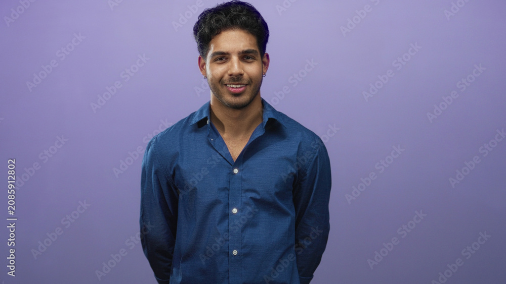Obraz premium Young man standing with hands behind back in studio, wearing a blue button shirt against a purple backdrop, eyes moving from closed to open with a slight smile; calm confidence.