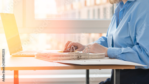 Professional person types on laptop at wooden desk. Warm sunlight streams through large window. City buildings visible outside. Focused demeanor suggests dedication