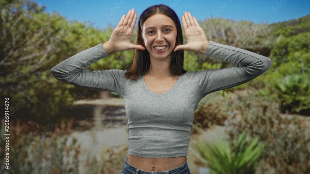 Fototapeta premium Young woman frames face with hands and smiles, visible bellybutton and midriff on a sunlit forest path; playfulness.