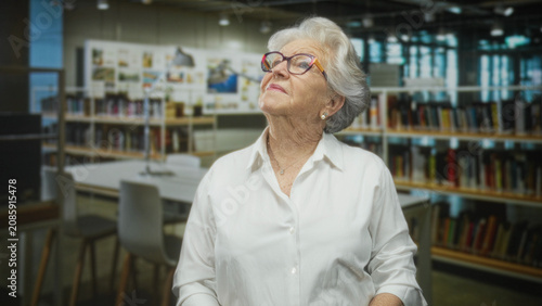 Foto Senior woman in white blouse wearing glasses looks up with chin lifted revealing necklace in a library building; quiet contemplation