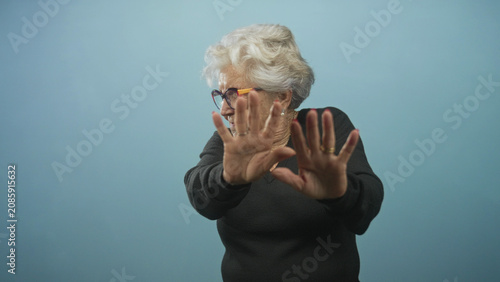 Foto Senior woman wearing glasses and rings holds hands palms out in a stop gesture in studio blue backdrop; defensive refusal