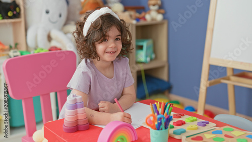 Fotografie Little girl smiling and drawing with a pencil at a colorful play table with crayons, wooden puzzle and toys next to an easel in a building; playful creativity