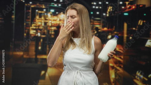 Принти на полотні Blonde woman holding milk bottle, hand to mouth covering mouth in a building with store shelves visible; disgust reaction