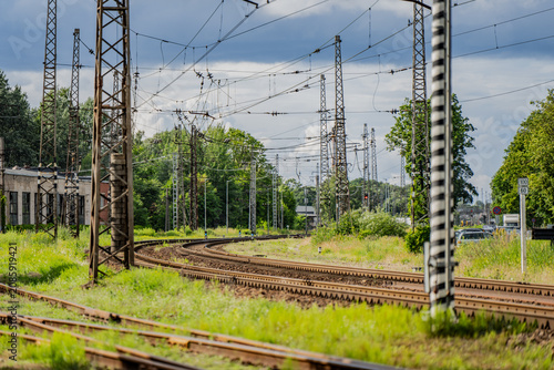 Industrial railway tracks curving through a green landscape with tall metal catenary masts, overhead power lines, and electrification infrastructure