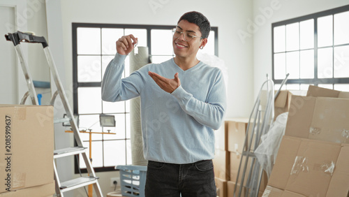 Photos Man holding house key between fingers and presenting it with open palm amid stacked moving boxes and ladder in bright building; joy new home