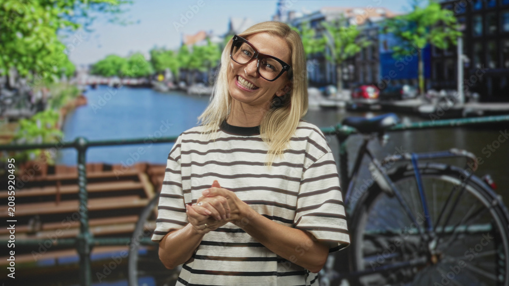 Fototapeta premium Woman with glasses clasping hands beside a bicycle on a studio canal backdrop, striped shirt and broad smile leaning toward camera, friendly expression and soft lighting; joyful warmth.