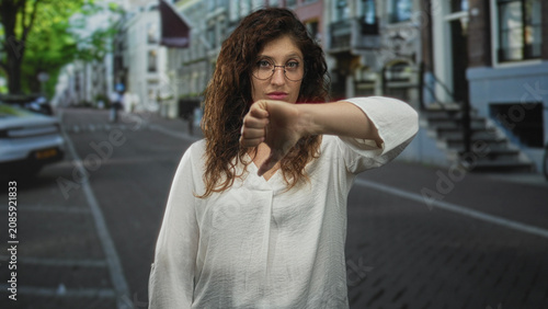 Εκτύπωση καμβά Woman wearing glasses shows thumbs down with her hand on a city street lined with buildings and parked cars; disapproval resolve