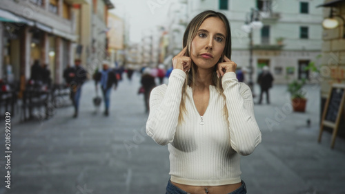 Obraz na plátně Woman with navel piercing, fingers in ears to block noise on a busy crowded city street; annoyance coping