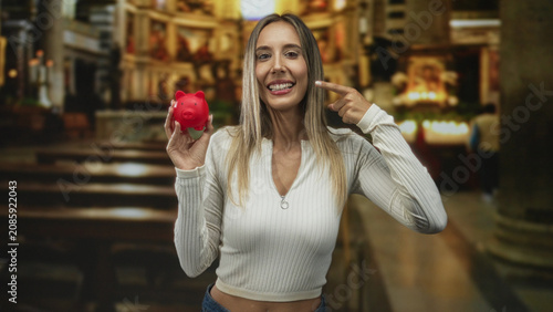 Billede på lærred Young woman holding red piggybank with fist pump gesture in church building; hope savings charity community