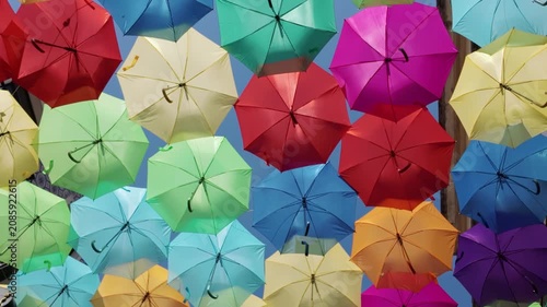 A dense arrangement of multi-colored umbrellas creates a festive overhead canopy. Sunlight filters through the fabric panels, highlighting saturated hues and soft shadows in a joyful urban installatio