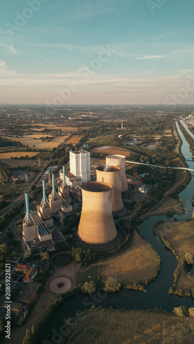 Industrial power plant with cooling towers surrounded by green fields and a canal under a clear blue sky, captured at sunset — symbol of modern energy and landscape harmony.