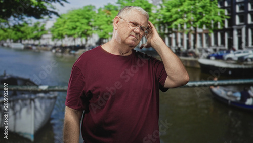 Bild auf Leinwand Man touching his temple with right hand by canal railing, wearing maroon t shirt and glasses, boats and narrow canal houses visible; pensive reflection
