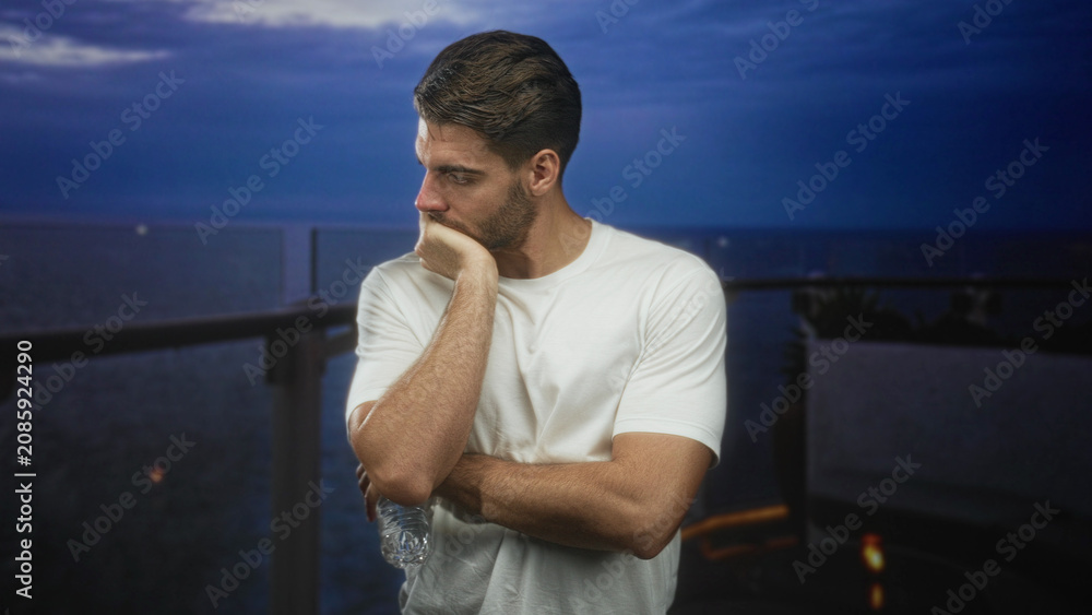Fototapeta premium Man with beard in white shirt holding water bottle, hand on chin leaning on balcony railing of building overlooking ocean at dusk; quiet contemplation.