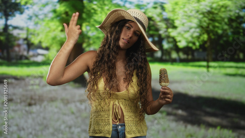 Fotografie Woman holding chocolate coated ice cream popsicle sprinkled with nuts, hand touching wide brim straw hat, wearing yellow knit camisole and denim jeans in sunlit park; summer serenity