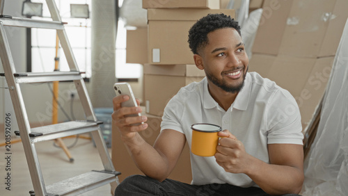 Foto Man holding smartphone and yellow mug, smiling while seated among stacked moving boxes in an indoor building; contentment moving
