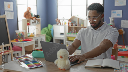 Schilderij op canvas Man wearing glasses with laptop, open notebook and plush chick on desk, arms crossed while seated among toys in a preschool classroom building; calm attentive teaching