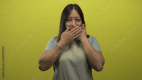 Bild auf Leinwand Woman covering mouth with both hands in studio against a yellow chartreuse backdrop wearing gray apron and short sleeve shirt; silence secrecy restraint