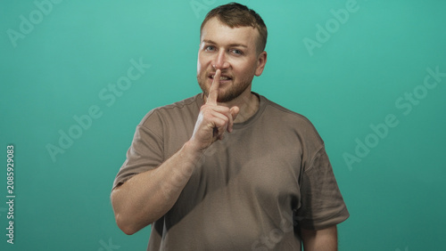 Canvastavla Man finger to lips for silence in teal studio, pointing gesture with visible fingertip and relaxed smile; secret quiet