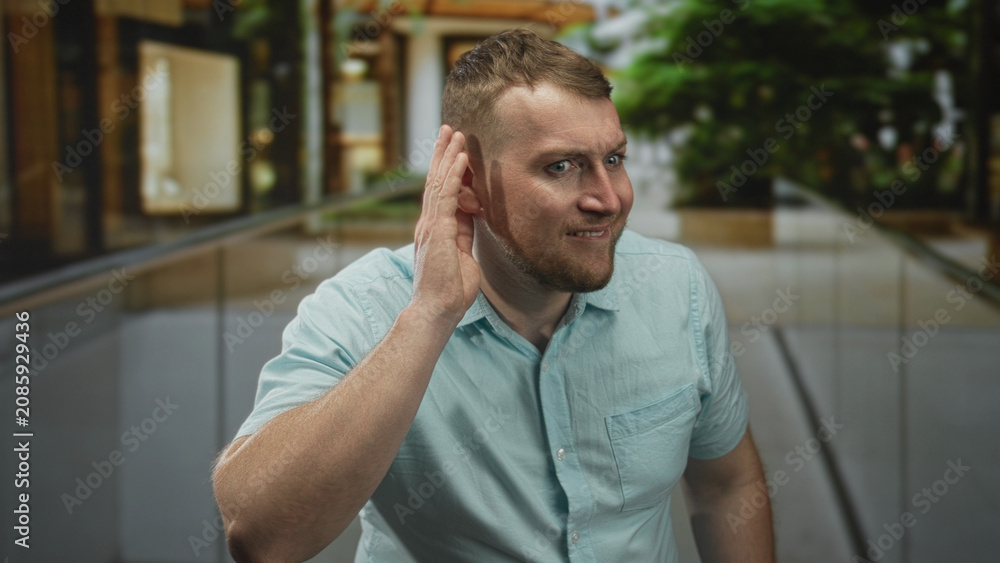 Fototapeta premium Man with hand cupped to ear leaning forward on indoor building walkway near glass railing and plants; curious listening communication.