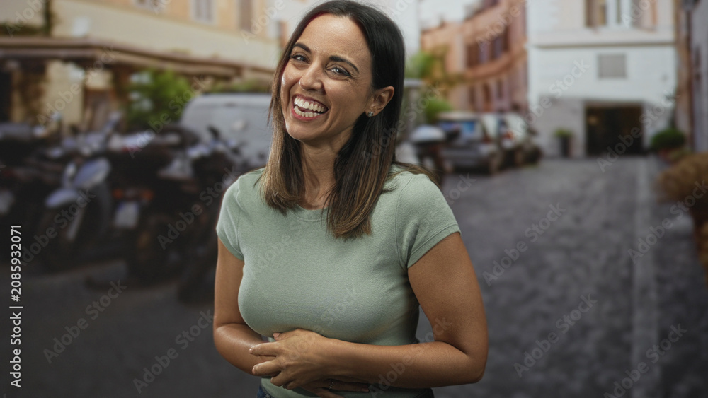 Fototapeta premium Woman laughing with hands clasped over her stomach on a street with parked motorcycles and cobblestone; joy warmth.