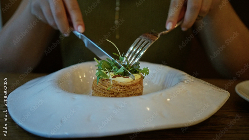 Fototapeta premium Woman cuts a layered pastry with knife and fork in building, close up on hands and plate; quiet mindful satisfaction.