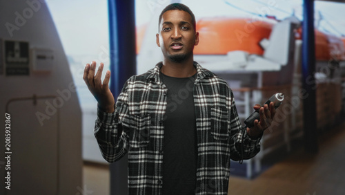 Fotografía Man holding electric screwdriver with right hand raised in questioning gesture in studio set with visible lifeboat backdrop; skill confidence