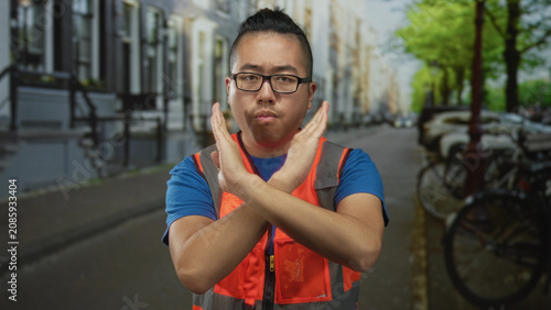 фотографія Man wearing orange reflective vest and blue shirt with glasses, arms crossed in front, standing on a city street lined with parked bicycles; rejection warning