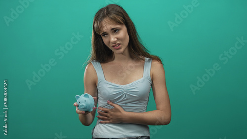 Bild auf Leinwand Young woman holding blue piggy bank and clutching stomach with one hand in teal studio; debt financial worry