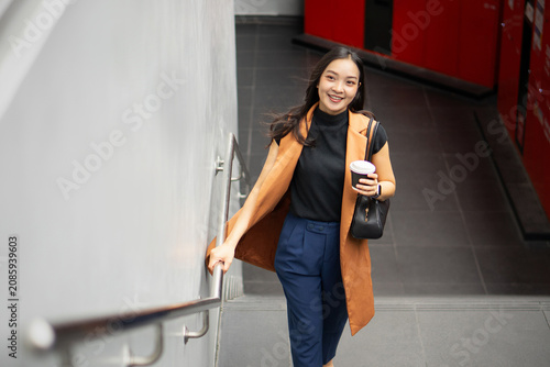 A stylish Asian woman smiles brightly while carrying a coffee cup and walking up an outdoor concrete staircase.