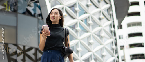 A bright Asian woman holds her smartphone and looks up thoughtfully while walking past a modern building exterior.