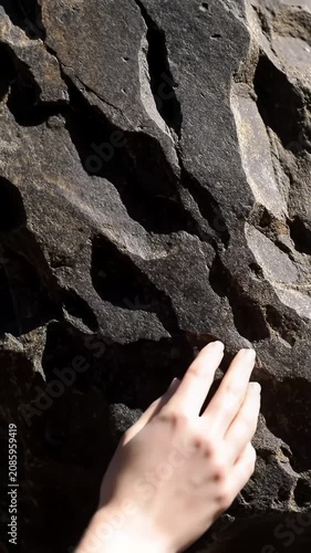 Extreme close-up video of a clean, neutral human hand's fingertips gently tracing a robust dark grey natural stone, highlighted by strong natural light. Trust and security concept