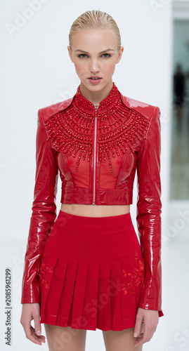 Stylish Female Model in Glossy Red Jacket and Pleated Skirt with Bold Embellishments, Captured in a Minimalist High-Fashion Studio Setting, Exuding Edgy Elegance and Confidence