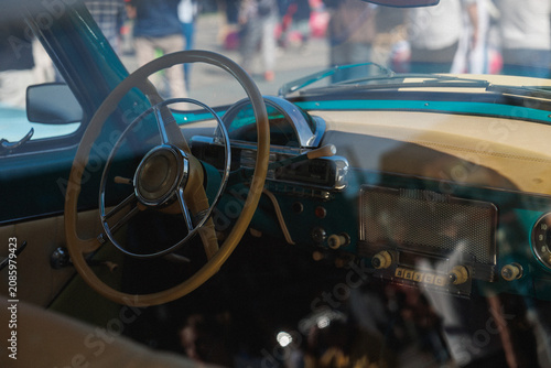 Close-up view through window of vintage turquoise car interior showing large circular steering wheel, chrome horn ring, retro dashboard with round gauges and pushbutton radio controls, cream leather