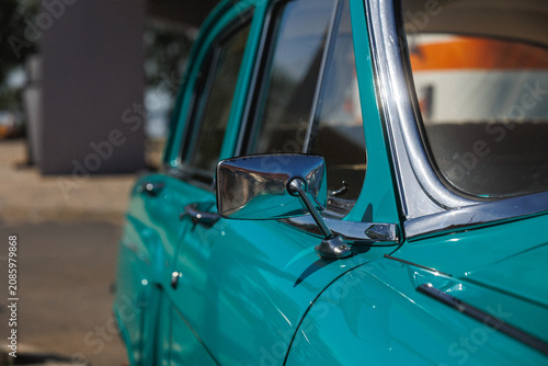 Closeup view of chrome side mirror mounted on teal painted classic car door, showing reflective trim, glossy finish, curved windshield edge, and bright sunlit surface detail, with car, classic