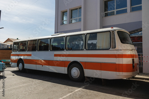Side view of retro orange and white coach bus parked in lot next to pale modern building with large windows; sunlight casts soft shadows across bus body and asphalt, interior rows of passenger seats