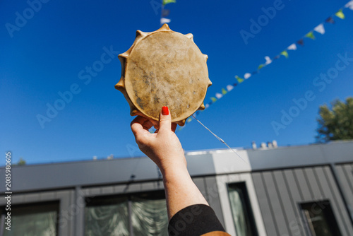 Close-up of hand with red nail polish holding round leather frame drum aloft against deep blue sky, while triangular bunting and modern building facade add festive urban context and bright daylight